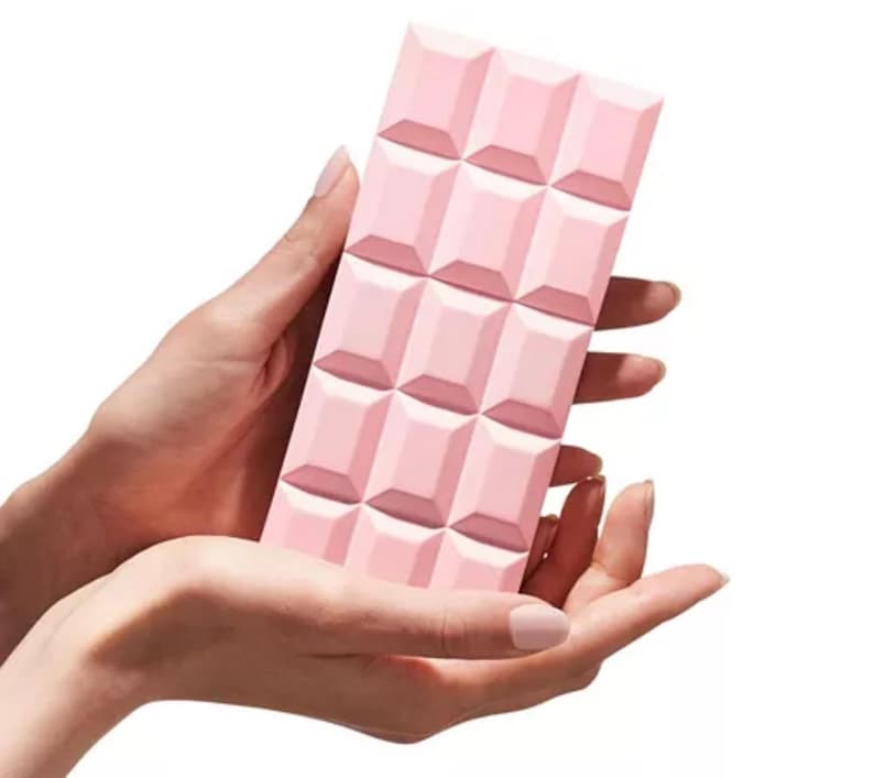 Puede incluir: Un objeto rectangular rosa con un patr&oacute;n de cuadr&iacute;cula, que se asemeja a una tableta de chocolate, se sostiene en dos manos. El objeto tiene un acabado mate y es de color rosa uniforme. Las manos son de piel clara y el fondo es blanco.