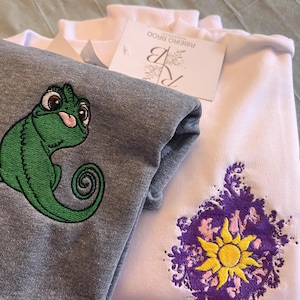 Peut inclure: Deux sweat-shirts pliés, l'un gris avec une broderie de caméléon vert, l'autre blanc avec un motif de soleil violet et jaune. Une étiquette Ribeiro Brood est visible. Les sweat-shirts sont empilés.