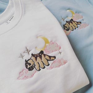 Pode incluir: Camisolas brancas e azul claro com desenhos bordados. Os desenhos apresentam uma criatura de desenho animado com uma sela, uma lua crescente amarela, nuvens rosa e estrelas. As camisolas são feitas de um material macio.