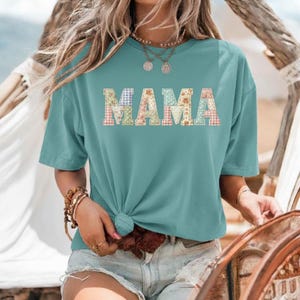 Puede incluir: Camiseta de color turquesa con la palabra "MAMA" en letras de patchwork. Las letras están hechas de varios patrones florales y de cuadros. La camiseta de manga corta está anudada a la cintura.