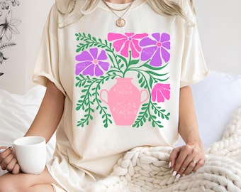 Puede incluir: Camiseta color crema con un jarrón rosa con flores moradas y rosas y hojas verdes. El diseño floral está centrado en la camiseta. La camiseta tiene mangas cortas y un corte holgado.