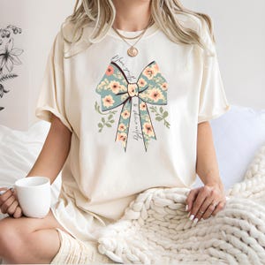 May include: Cream-coloured t-shirt featuring a large bow design with floral patterns in peach and blue. The bow has the text "Bloom gently & shine wildly" printed on it. The shirt is a casual style.