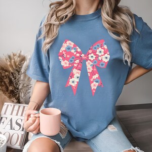 May include: A blue t-shirt featuring a large pink bow with a floral pattern of white and blue flowers. The shirt has short sleeves and a relaxed fit. The bow design is centered on the front of the shirt.