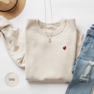 Może przedstawiać: Kremowa bluza z małą, czerwoną biedronką wyhaftowaną na piersi. Srebrny naszyjnik z wisiorkiem spoczywa na bluzie. Słowo "SAND" jest wydrukowane na małej, okrągłej, kremowej etykiecie. Na zdjęciu znajduje się również słomkowy kapelusz i dżinsy.