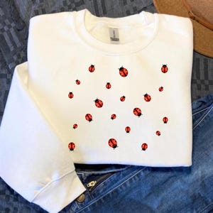 May include: White crew neck sweatshirt decorated with a pattern of red ladybugs with black spots. The long-sleeved top is made of a soft material. The sweatshirt is shown with blue jeans.
