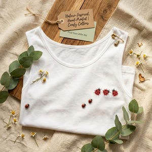 May include: A white vest top featuring a ladybug pattern. The garment is adorned with red ladybug illustrations. The tag reads "Nature-Inspired, Hand-Printed, Comfy Cottons" and "Find Your Little Joy".
