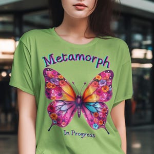 Könnte beinhalten: Ein grünes T-Shirt mit einem bunten Schmetterlingsmotiv und dem Text "Metamorph In Progress".