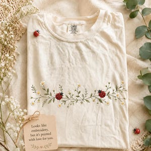 Könnte beinhalten: Ein cremefarbenes T-Shirt mit einem horizontalen Aufdruck aus grünen Ranken, kleinen weißen Blumen und zwei roten Marienkäfern auf der Brust. Ein kleiner roter Marienkäfer ist in der Nähe des Halsausschnitts aufgedruckt. Das Etikett zeigt Comfort Colors. Ein Schild sagt: Looks like embroidery, but it's printed with love for you.