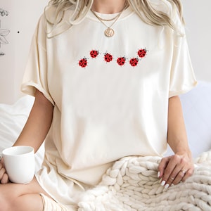 Könnte beinhalten: Cremefarbenes T-Shirt mit einer Reihe von sieben roten Marienkäfer-Designs. Die Marienkäfer haben schwarze Punkte und sind in einer gebogenen Linie angeordnet. Das Shirt hat kurze Ärmel und eine lockere Passform.