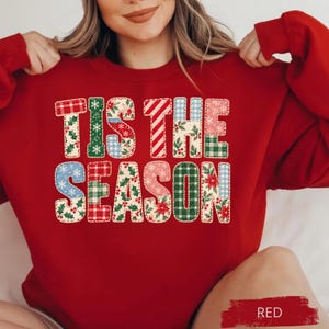 May include: A red crewneck sweatshirt featuring the text TIS THE SEASON in large block letters. Each letter is filled with a variety of holiday-themed patterns including red and green plaid, holly leaves, poinsettias, and snowflakes.