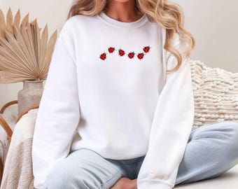 Puede incluir: Sudadera blanca con una línea de siete diseños de mariquitas rojas. Cada mariquita tiene puntos negros y está dispuesta en una línea curva. La sudadera es de color blanco liso.