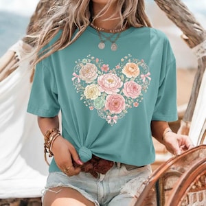 Könnte beinhalten: Türkisfarbenes T-Shirt mit einem floralen Herzdesign. Das Herz besteht aus pastellfarbenen Blumen, darunter rosa, gelbe und pfirsichfarbene Rosen, mit zarten grünen Blättern und kleinen rosa Schleifen. Das T-Shirt ist an der Taille geknotet.