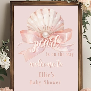 May include: A framed baby shower sign with a light pink background. The sign features a seashell, a pink ribbon, and the words "little pearl is on the way, welcome to Ellie's Baby Shower."