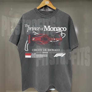 May include: A dark gray t-shirt with a faded vintage look featuring a red Formula 1 race car on the front. The text "The Prince of Monaco" and "Circuit de Monaco" are printed on the shirt. The shirt also features the Formula 1 logo.