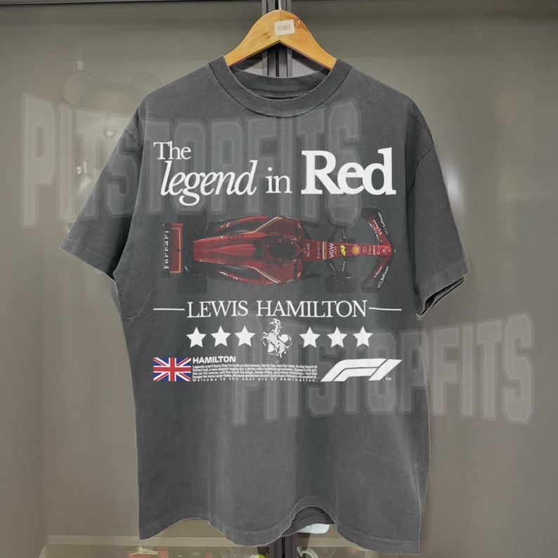 Puede incluir: Una camiseta negra con un estampado descolorido de un coche de carreras rojo y el texto "The legend in Red - Lewis Hamilton". La camiseta tambi&eacute;n presenta una bandera del Reino Unido y el logotipo de la F&oacute;rmula 1.