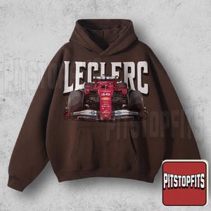 May include: A brown hooded sweatshirt featuring a red race car graphic with the number 16. The word "LECLERC" is printed in white across the top. The sweatshirt has a front pocket and a drawstring hood. The Pitstopfits logo is in the bottom right corner.