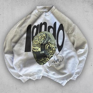 May include: A light gray sweatshirt with the word "Lando" in large black letters. A racing helmet with a black visor and a yellow and black pattern is in front of the sweatshirt. The helmet has the Monster Energy logo.