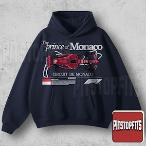 Peut inclure: Sweat à capuche bleu marine avec un dessin de voiture de course rouge et le texte "The Prince of Monaco". Le design comprend "Circuit de Monaco" et "2:23:15". Un logo F1 blanc est également visible.