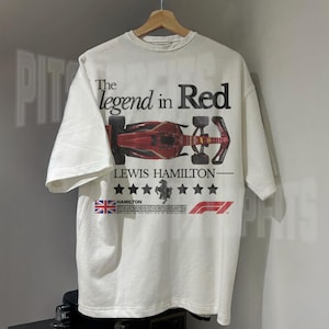 Puede incluir: Camiseta blanca con un gr&aacute;fico de coche de carreras de F&oacute;rmula 1 rojo, negro y blanco. El texto "The legend in Red" est&aacute; por encima del coche y "Lewis Hamilton" est&aacute; por debajo del coche. El texto "Hamilton" est&aacute; en una fuente m&aacute;s peque&ntilde;a y est&aacute; debajo de una bandera del Reino Unido.