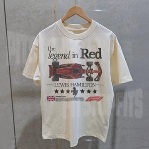 Puede incluir: Una camiseta blanca con un gr&aacute;fico de coche de carreras de F&oacute;rmula 1 rojo, negro y blanco. El texto "The legend in Red - Lewis Hamilton" est&aacute; impreso en la camiseta. La camiseta tambi&eacute;n presenta una bandera del Reino Unido y el logotipo de la F&oacute;rmula 1.
