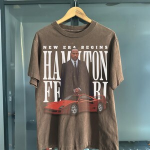 Peut inclure: Un t-shirt marron &agrave; l'aspect d&eacute;lav&eacute; avec un imprim&eacute; d'un homme en costume debout &agrave; c&ocirc;t&eacute; d'une Ferrari rouge avec le texte "NEW ERA BEGINS HAMILTON FERRI" imprim&eacute; sur le devant.