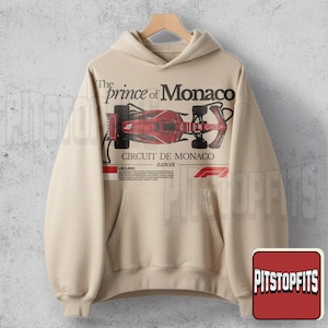 May include: Beige hoodie featuring a red and black Formula 1 race car graphic. The text "The Prince of Monaco" and "Circuit de Monaco" are printed above and below the car illustration. The hoodie has a front pocket and a drawstring hood.