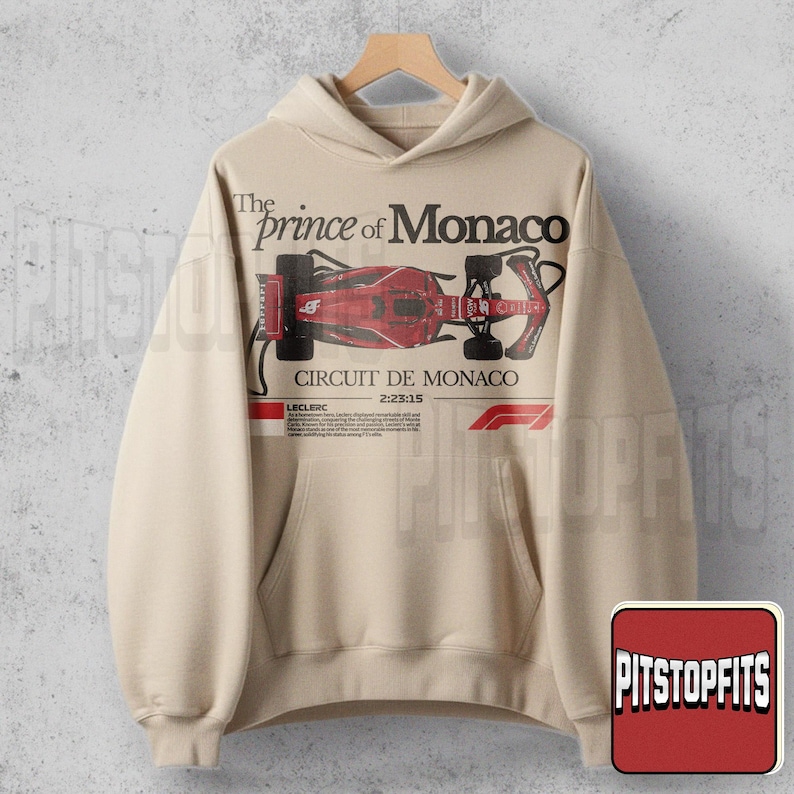 May include: Beige hoodie featuring a red and black Formula 1 race car graphic. The text "The Prince of Monaco" and "Circuit de Monaco" are printed above and below the car illustration. The hoodie has a front pocket and a drawstring hood.