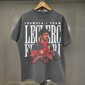May include: A black t-shirt with a graphic print of a Formula 1 race car driver. The text "FORMULA 1 TEAM LECLERC FERRARI" is printed on the shirt.