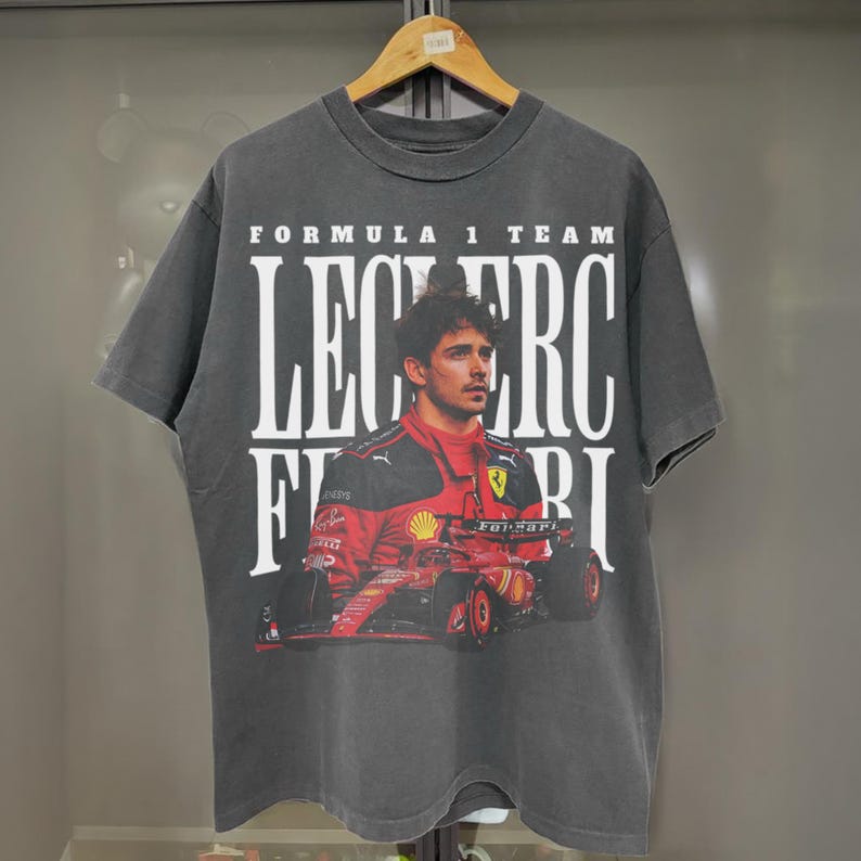 May include: A black t-shirt with a graphic print of a Formula 1 race car driver. The text "FORMULA 1 TEAM LECLERC FERRARI" is printed on the shirt.