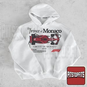 May include: White hoodie featuring a red race car graphic with the text "The Prince of Monaco" and "Circuit de Monaco 2:23:15". The design includes the F1 logo and a small text box with the name "Leclerc".