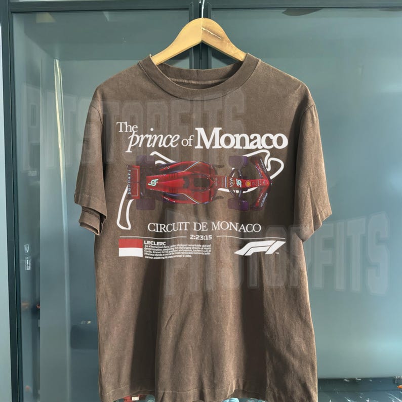 May include: Brown t-shirt with a faded vintage look featuring a graphic of a Formula 1 race car on the Circuit de Monaco. The text "The prince of Monaco" is printed above the car graphic.