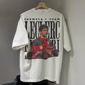 May include: White t-shirt with a graphic of Formula 1 driver Charles Leclerc in a red Ferrari race car. The text "Formula 1 Team Leclerc Ferrari" is printed on the shirt.