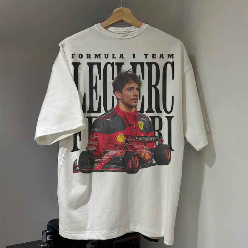 May include: White t-shirt with a graphic of Formula 1 driver Charles Leclerc in a red Ferrari race car. The text "Formula 1 Team Leclerc Ferrari" is printed on the shirt.