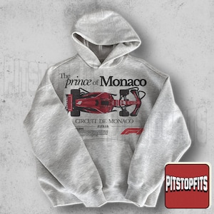 May include: A light gray hoodie with a hood and a front pocket. The front of the hoodie features a red and black graphic of a race car with the text "The Prince of Monaco" and "Circuit De Monaco".
