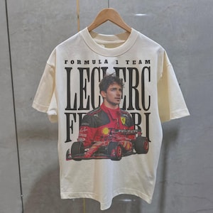 May include: A white t-shirt with a black and red graphic print of a Formula 1 race car driver, Charles Leclerc, with the text "Formula 1 Team Leclerc Ferrari".
