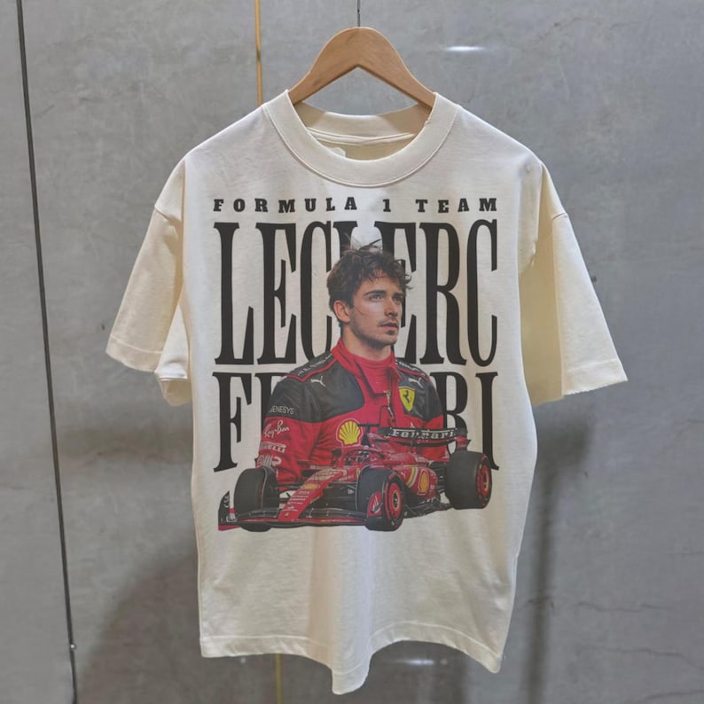 May include: A white t-shirt with a black and red graphic print of a Formula 1 race car driver, Charles Leclerc, with the text "Formula 1 Team Leclerc Ferrari".
