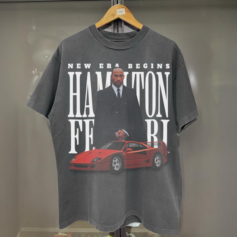 Peut inclure: Un t-shirt noir avec un imprim&eacute; graphique d'un homme en costume debout &agrave; c&ocirc;t&eacute; d'une Ferrari rouge. Le texte "NEW ERA BEGINS HAMILTON FERRARI" est imprim&eacute; sur le t-shirt.