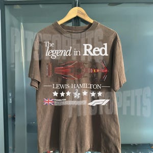 Puede incluir: Una camiseta marr&oacute;n con un estampado gr&aacute;fico blanco de un coche de carreras de F&oacute;rmula 1. El texto "The legend in Red - Lewis Hamilton" est&aacute; impreso en la camiseta. La camiseta tambi&eacute;n presenta una bandera del Reino Unido y el logotipo de la F&oacute;rmula 1.