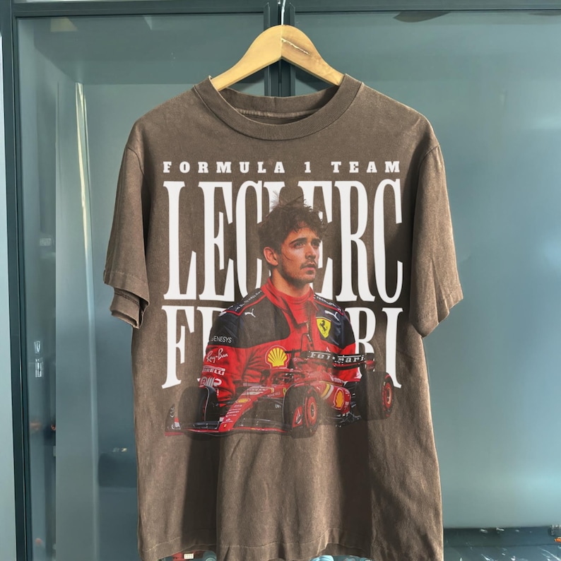 May include: A brown t-shirt with a graphic print of a Formula 1 race car driver, Charles Leclerc, in a red Ferrari race car. The text "Formula 1 Team Leclerc Ferrari" is printed on the shirt.