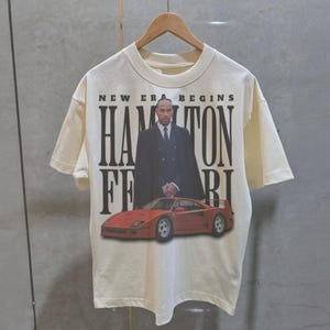 Peut inclure: Un t-shirt blanc avec un imprim&eacute; noir d'un homme en costume debout &agrave; c&ocirc;t&eacute; d'une Ferrari rouge. Le texte sur le t-shirt est "New Era Begins Hamilton Ferrari".