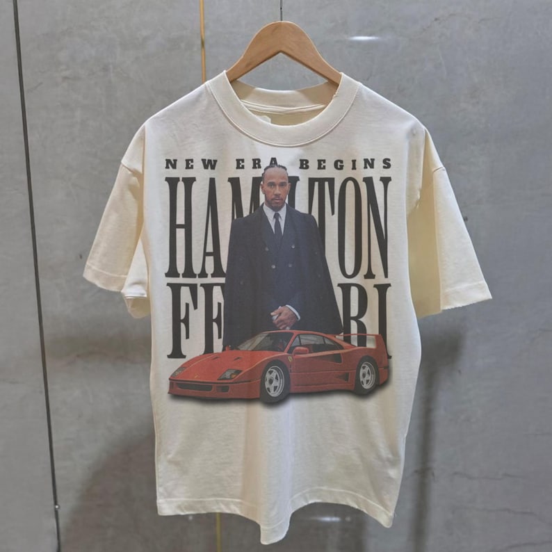 Peut inclure: Un t-shirt blanc avec un imprim&eacute; noir d'un homme en costume debout &agrave; c&ocirc;t&eacute; d'une Ferrari rouge. Le texte sur le t-shirt est "New Era Begins Hamilton Ferrari".