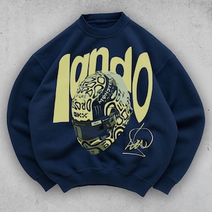 May include: Navy blue sweatshirt featuring the name "lando" in a yellow, bold font. The design includes a detailed illustration of a racing helmet with various logos and a signature. The sweatshirt is a crew neck style.