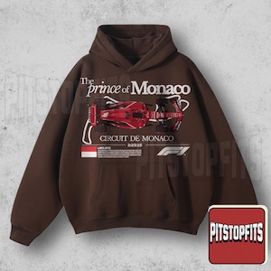 May include: Brown hoodie with a front graphic featuring a red race car and the text "The Prince of Monaco CIRCUIT DE MONACO". The hoodie has a kangaroo pocket and a drawstring hood.