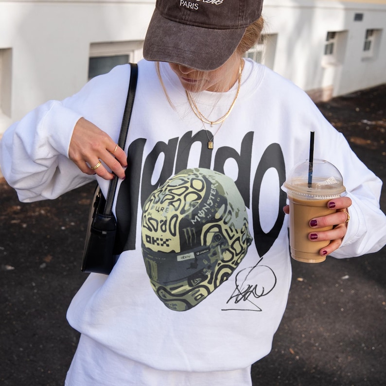May include: White sweatshirt with the word "Lando" in large black letters. The sweatshirt features a graphic of a racing helmet with a camouflage design and the words "Monster Energy" and "OKX". A brown cap and a black purse are also visible.