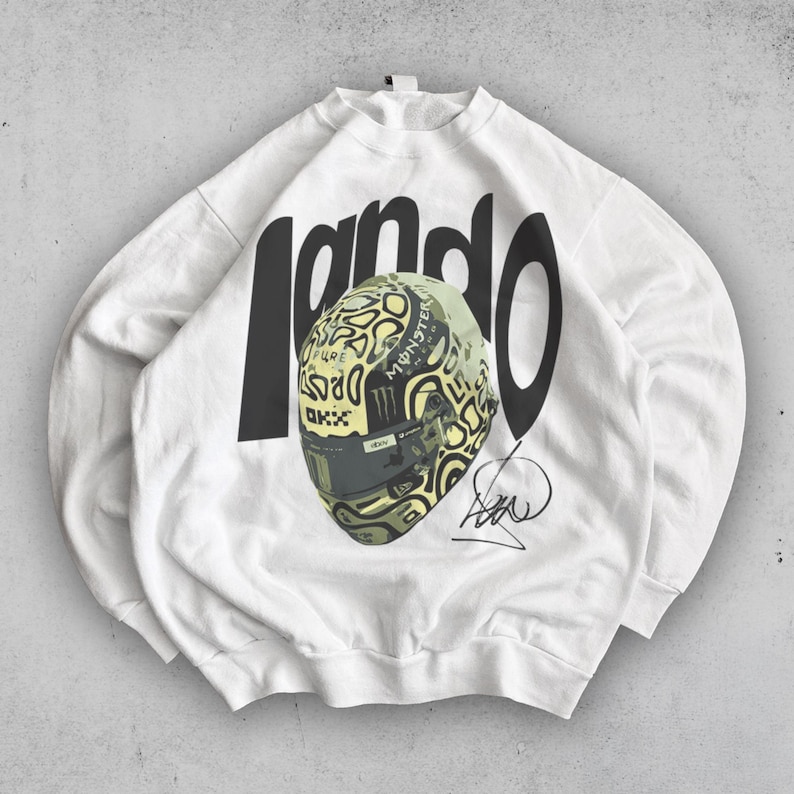 May include: White sweatshirt with the word "Lando" in large black letters. A racing helmet with a camouflage pattern and the Monster Energy logo is in the center. A signature is below the helmet.