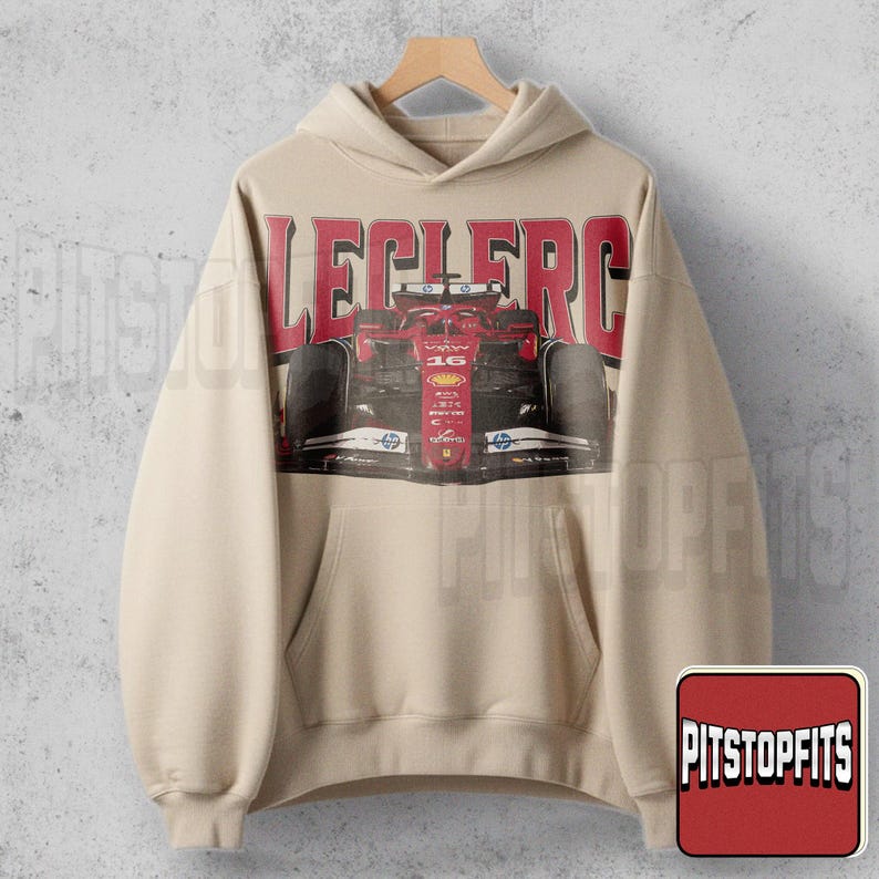 May include: Beige hoodie with a front graphic of a red race car and the name "LECLERC" in red block letters. The hoodie has a kangaroo pocket and a drawstring hood. The image is of a Formula 1 race car.