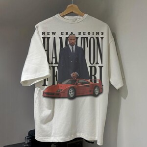 Peut inclure: T-shirt blanc avec un motif rouge et noir d'une voiture de course de Formule 1 et d'un homme en costume. Le texte "New Era Begins Hamilton Ferrari" est imprim&eacute; sur le t-shirt.