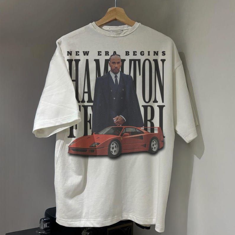 Peut inclure: T-shirt blanc avec un motif rouge et noir d'une voiture de course de Formule 1 et d'un homme en costume. Le texte "New Era Begins Hamilton Ferrari" est imprim&eacute; sur le t-shirt.