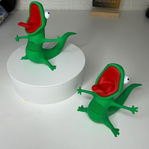 May include: Two green lizard figurines with wide-open red mouths, protruding tongues, and large white eyes. The figures have articulated, ribbed midsections and outstretched limbs. A small sign in the background displays the text PARS3D STUDIO.