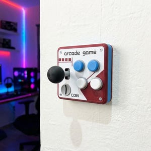 Può includere: Un controller per giochi arcade rettangolare montato a parete con un joystick nero, due pulsanti blu, due pulsanti bianchi e una fessura per monete. La piastra frontale è bianca con accenti rossi e il testo arcade game e COIN in nero.